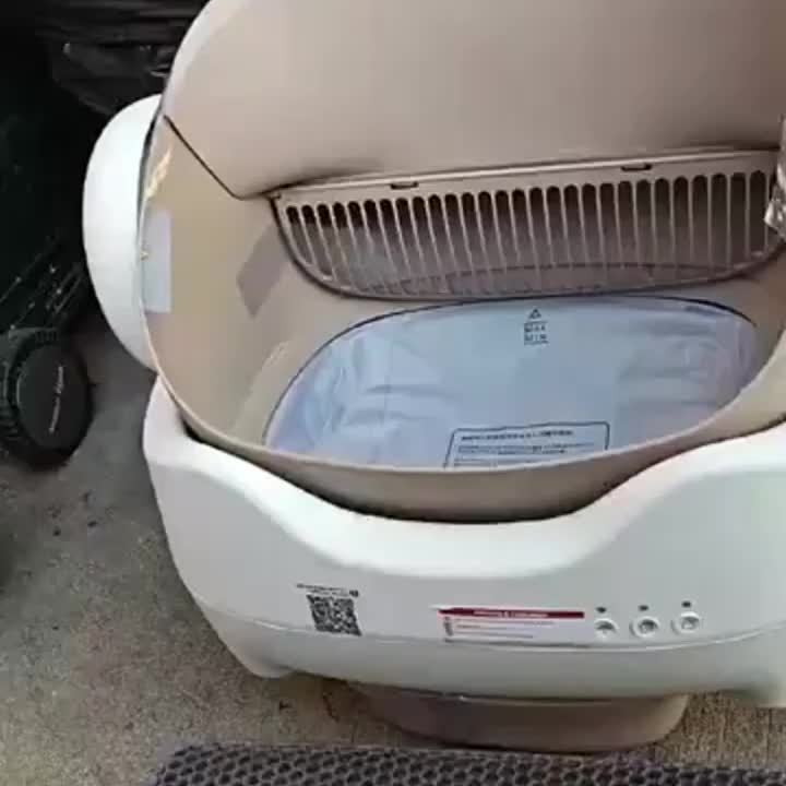 Beige and white automatic cat litter box, possibly a dryer or washer, with visible branding on a concrete floor.