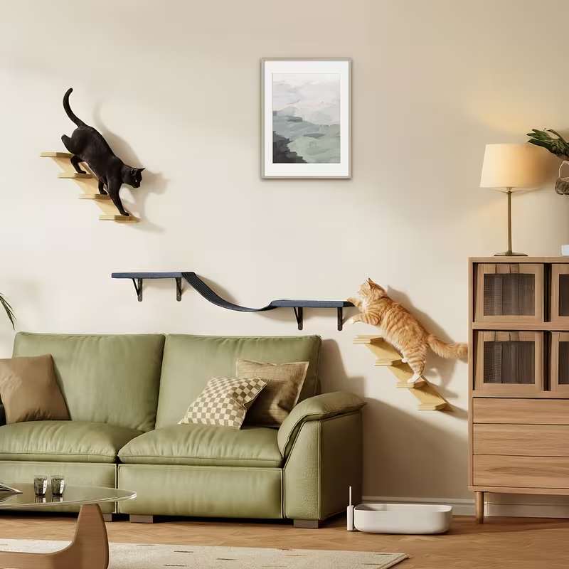 A styled living room with a green sofa, wall-mounted cat steps, a black cat climbing a wall cat tree, an orange tabby cat on a floor cat tree, and framed wall art above the couch.