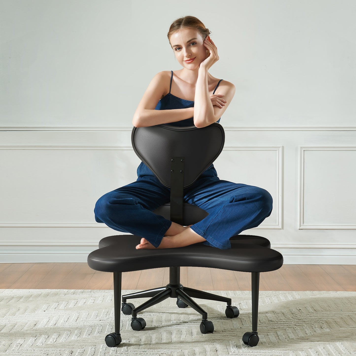 Woman sitting on a black office chair with a footrest in a room with white walls.