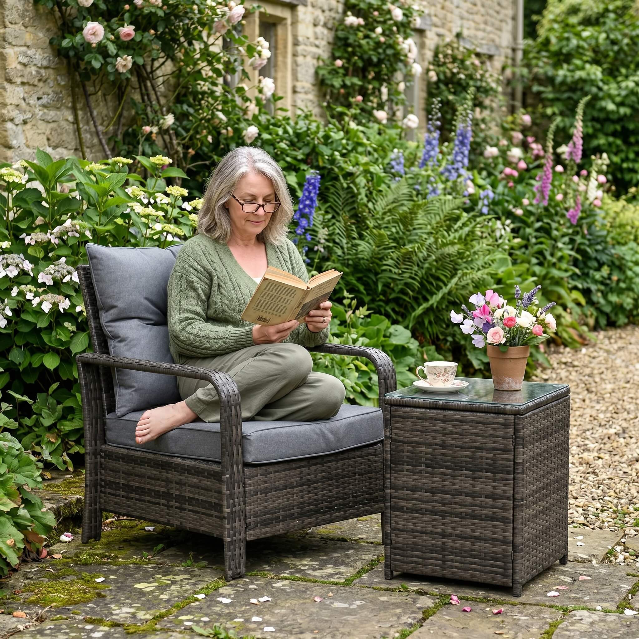 Woman reading a book in a garden setting with flowers and a small table.