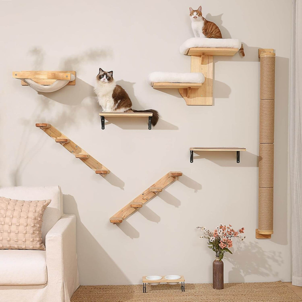 A wall-mounted cat tree set with multiple wooden platforms, steps, a scratching post, and cushioned perches installed on a light beige wall in a styled room. Two cats are sitting on the different platforms, and a cream sofa, pillow, and small flower vase are visible in the room.