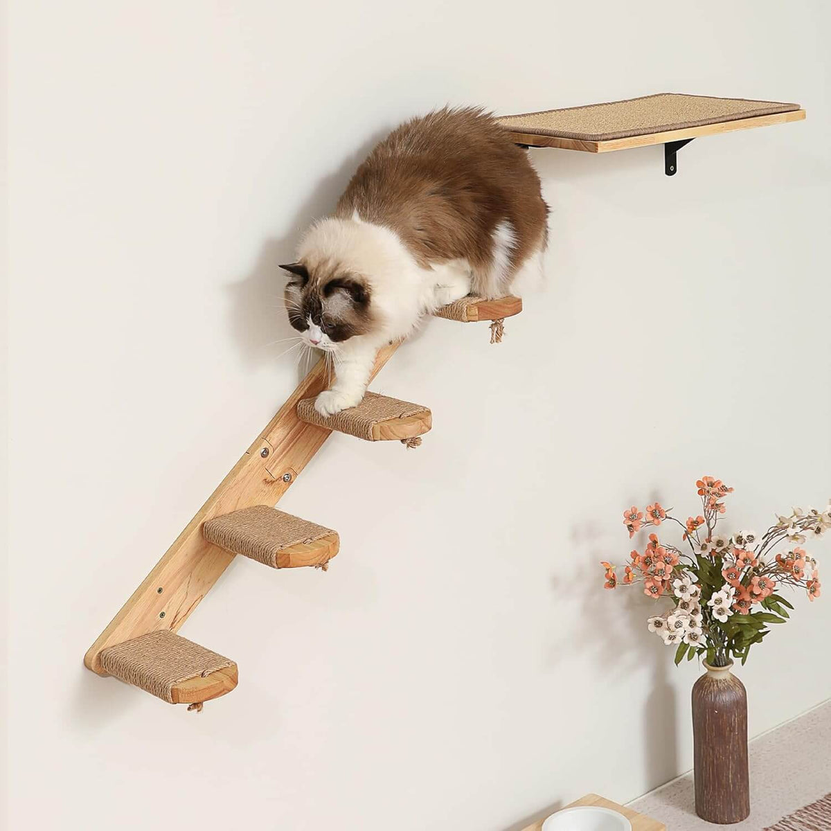 A fluffy brown and white cat is climbing a wooden wall-mounted cat tree with sisal-wrapped steps and a top perch against a plain light wall. A vase of flowers sits on the floor nearby.