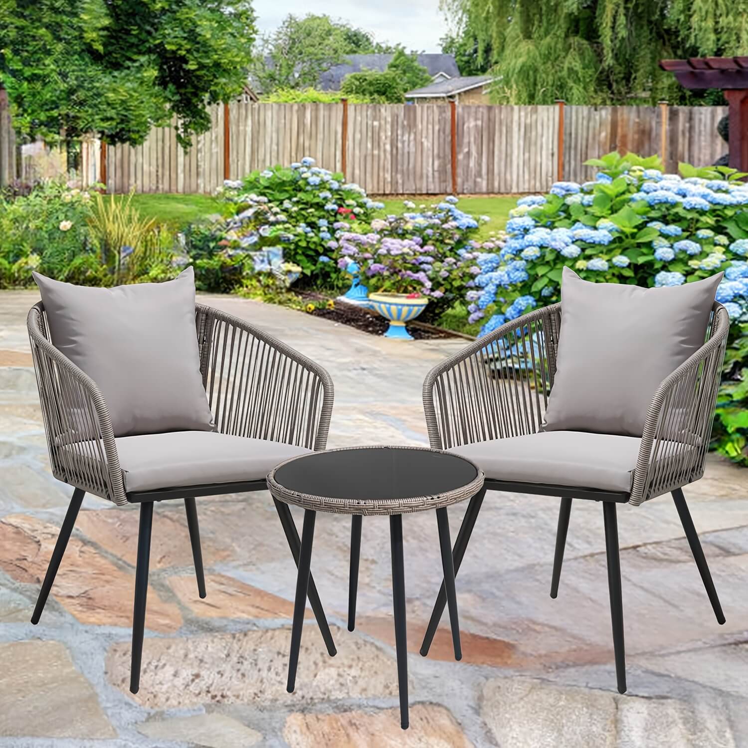 Outdoor patio set with two chairs and a table on a stone patio with flowers and trees in the background.