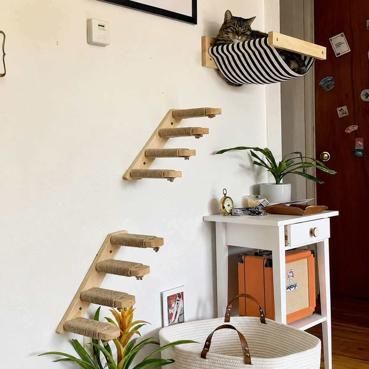 A styled interior corner showing wall-mounted cat climbing stairs and a striped cat bed with a cat resting inside, alongside a small white side table, plants, and home decor.
