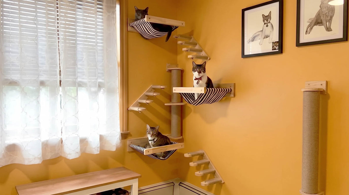 A multi-level cat tree with wooden stairs and striped fabric platforms in a home setting, with three cats resting on the different levels against a yellow wall.