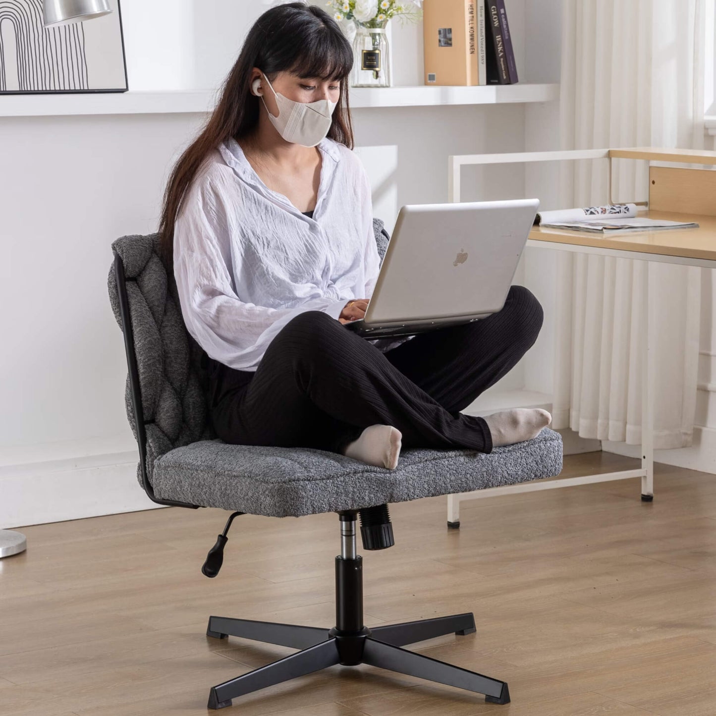 A woman working on her laptop while wearing a face mask and sitting cross legged on a fabric office chair