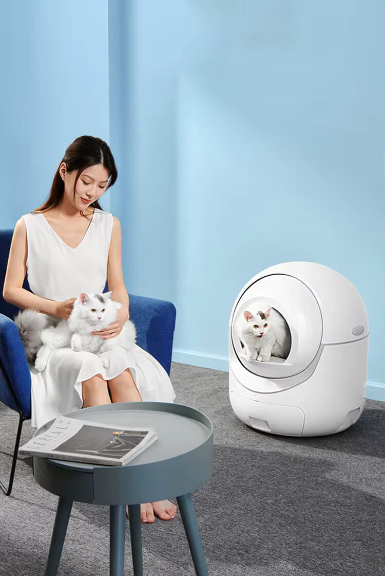 Woman with a cat next to a white self cleaning cat litter box in a room with light blue walls.