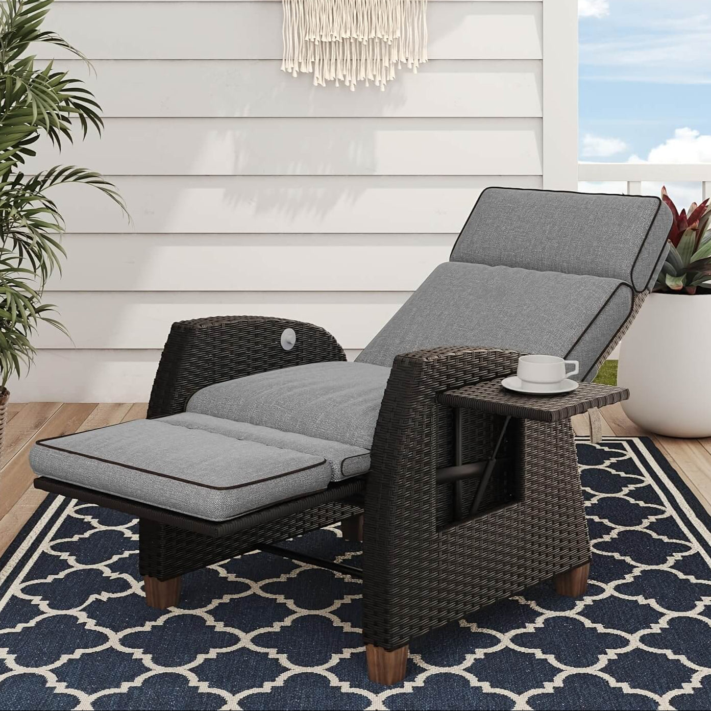 Outdoor lounge chair with gray cushions on a patio with plants and a rug.