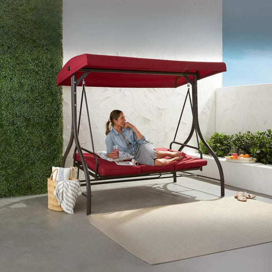 Person relaxing on a red outdoor porch swing set  with canopy in a garden setting.