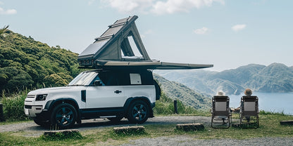 White Land Rover Defender with an open roof tent parked by a scenic lakeside.