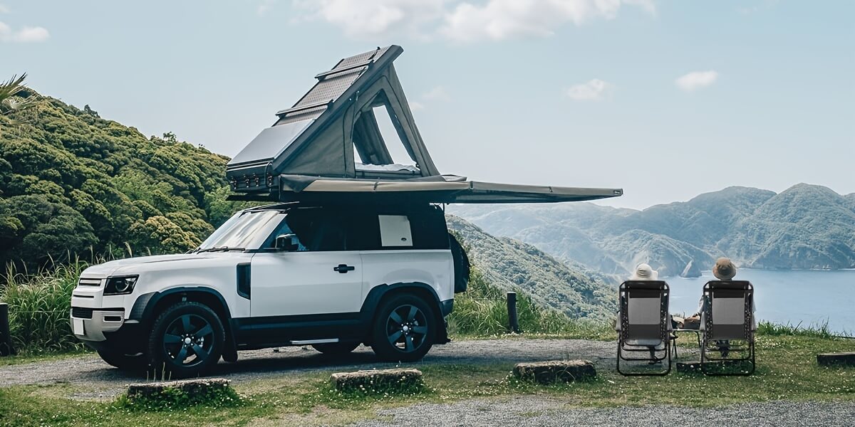 White Land Rover Defender with an extended roof tent parked by a scenic lakeside and a couple sitting on outdoor patio recliners beside it.