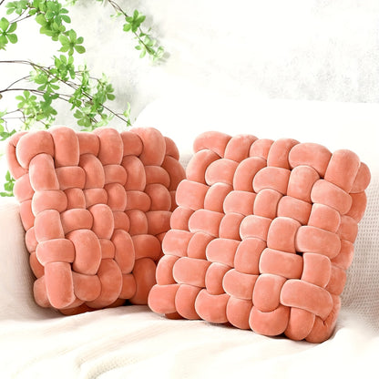 Two pink woven pillows on a white surface with a blurred green plant in the background